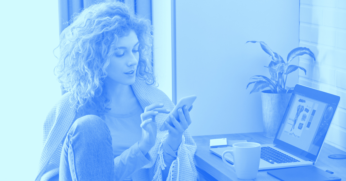 Woman looking at her phone while her laptop is open in front of her.